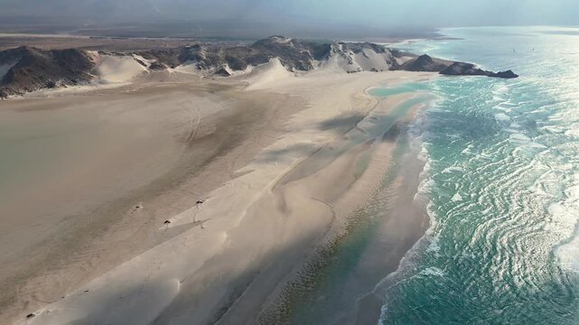 Socotra island, Aerial view of a vast sandy coastline where desert dunes and rugged mountains meet turquoise ocean lagoon waters, with dramatic shadows and bright sunlight reflecting across the sea.