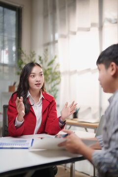 Young businesswoman in a red jacket explaining strategy and gesturing across a desk while consulting with a male client in a modern office during an important meeting