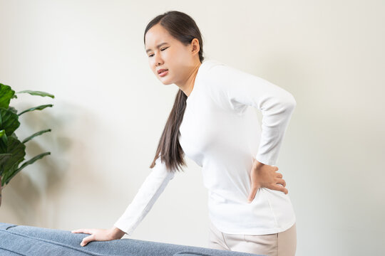 Asian young woman suffering from severe lower back pain and lumbar muscle injury while standing at home, backache and office syndrome concept for medical and health care person.