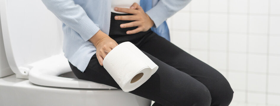 Stomach ache, sick asian young woman holding stomach in pain and hold tissue paper roll in bathroom sitting on toilet bowl, girl suffer from diarrhea her stomach painful. Hygiene, health care concept.