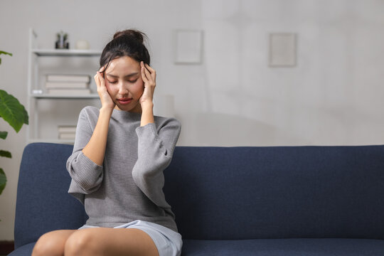 asian woman holding her temples while sitting on sofa at home with pain and stress, copy space for healthcare design. headache, migraine, pain, stress, health. Headache and migraine concept.