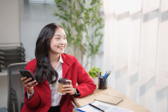 Professional Asian businesswoman smiling, looking away and feeling inspired while holding a smartphone and coffee cup in a modern office, planning future success