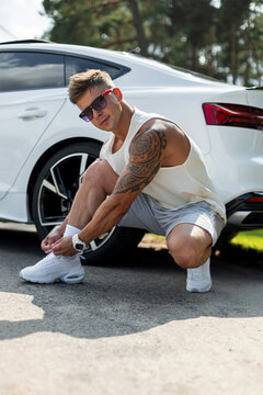 Muscular tattooed man in sunglasses crouching to tie his white sneakers beside a white luxury car, looking at camera. Athletic guy preparing for an outdoor workout in sunny nature.