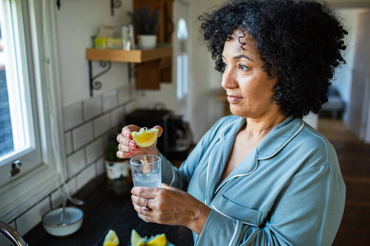 Woman in pajamas squeezing lemon into water at home kitchen