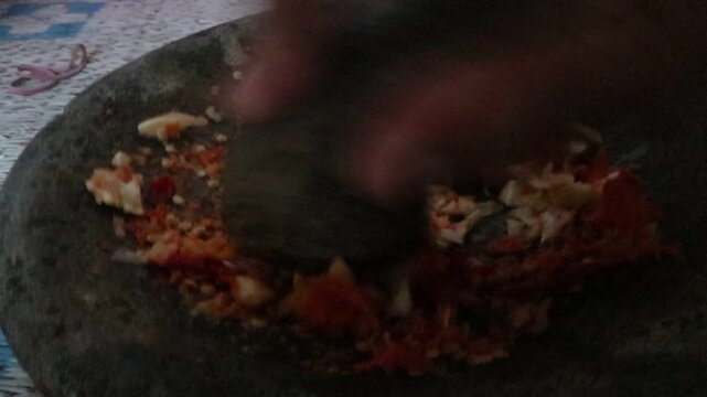 Rural Javanese housewife pounding and grinding garlic after chili in a traditional stone mortar to prepare homemade cooking spices in a village kitchen.