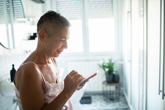 Smiling mature woman using smartphone in bright home bathroom