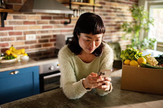 Woman using smartphone in home kitchen with fresh grocery delivery