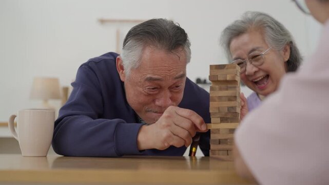 Asian senior man with friends playing wooden block tower game with concentration during indoor leisure activity. Active retirement lifestyle and wellness concept	