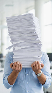A man in a blue shirt holds an overwhelming, precarious stack of white paper sheets. The feeling of being buried in paperwork, bureaucracy, or an excessive workload.