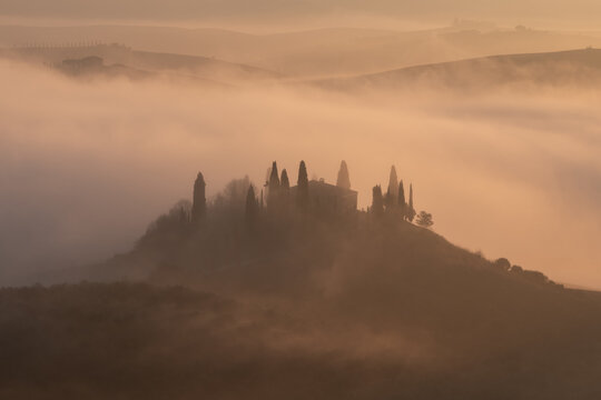 Aerial view of a traditional Tuscan villa surrounded by cypress trees during a misty sunrise, offering a peaceful and atmospheric rural landscape for travel and wellness.
