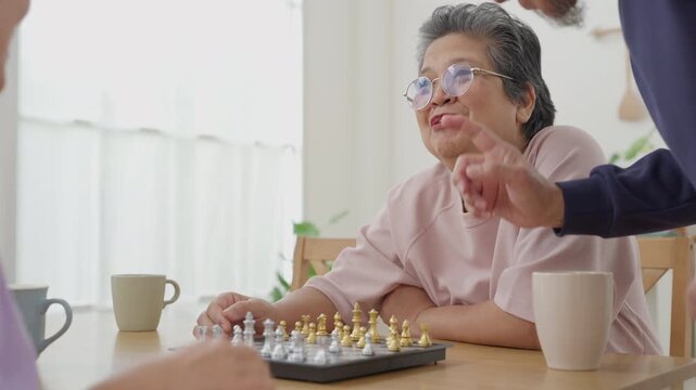 Asian senior women friends playing chess together with man watching during indoor leisure activity. Retirement lifestyle and cognitive wellness concept