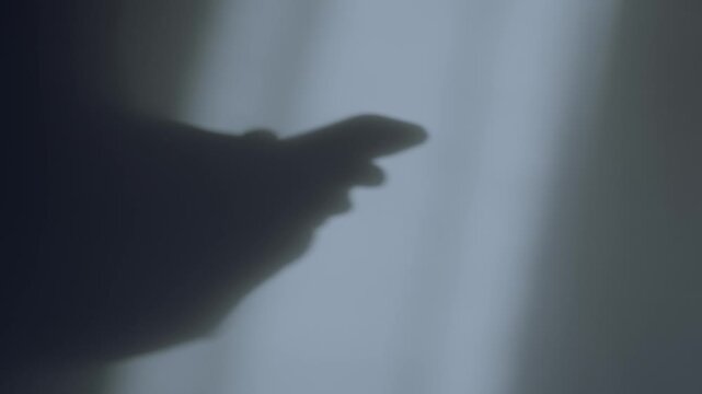 Silhouette shadow of person's hands typing and texting on mobile phone at night. Moonlight shining into office or room wall. Businessman or mysterious hacker or spy using smartphone to send messages