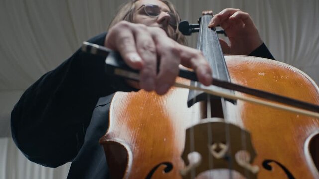 Caucasian young adult man with glasses playing cello, moving bow across strings in low angle shot. Ideal for classical music concert promotions, arts education videos, or cultural documentaries