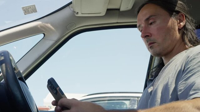 Driver in parked car focused on smartphone with low-angle close framing under daylight, with driver, phone, smartphone, man detail, showing car, interior, steering, hand context