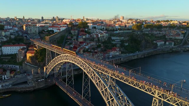 Drone view of Dom Luis I Bridge over Douro River in Porto, Portugal, with the river bend, including a tram on the bridge and rabelo boats by the quay at sunset. Orbit parallax shot