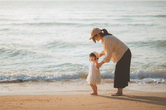 Mother helping toddler girl stand safely by the sea on sandy beach, caring support, childhood exploration, family vacation, summer travel, ocean experience and outdoor bonding lifestyle