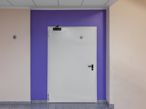 Single leaf white metal fire exit door with black handle and automatic closer hydraulic system installed on a purple painted wall section in a modern building hallway or public office or hospital