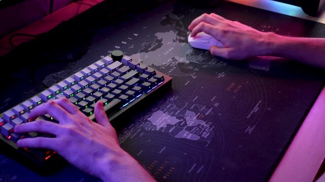 Close-up of a person using an RGB mechanical keyboard and computer mouse at a gaming desk. Colorful ambient lighting creates a modern immersive atmosphere while the hands operate the gaming controls.