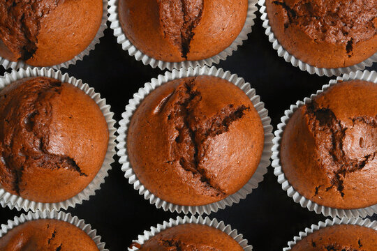 A repeating pattern layout of domestic chocolate muffins with cracked tops cooling down in white paper baking cups on a dark background.