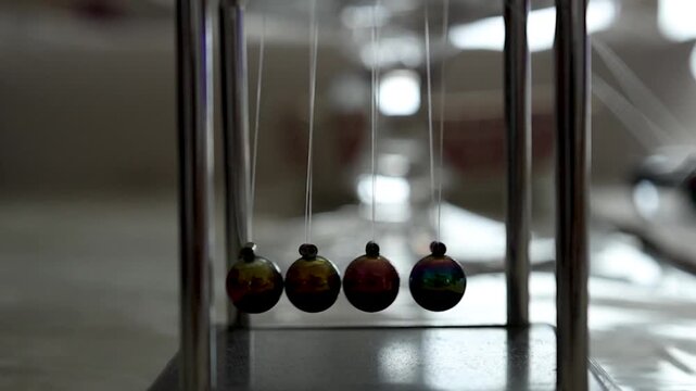 Wide shot of a Newton&rsquo;s cradle swinging in slow motion indoors. The reflective metallic balls move back and forth, illustrating momentum, balance and energy transfer in a classic physics desk toy.