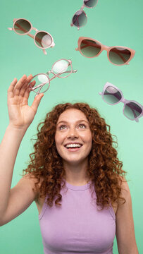 Happy young Caucasian woman with curly red hair smiling and catching different flying glasses against a light green background. Fun summer fashion, eyewear collection and youth lifestyle concept.