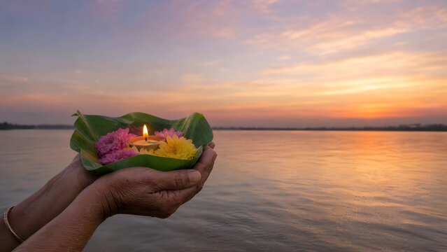 Hands holding burning diya and flowers over holy river water