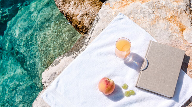 Relaxing summer vacation by the sea with a book, fresh fruit juice, peach, and white towel on coastal rocks