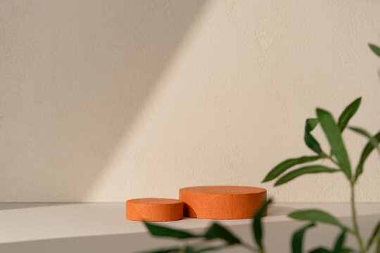 Two terracotta textured cylinder product display podiums on a white surface against a beige wall with diagonal light shadow, suitable for natural cosmetic product staging.