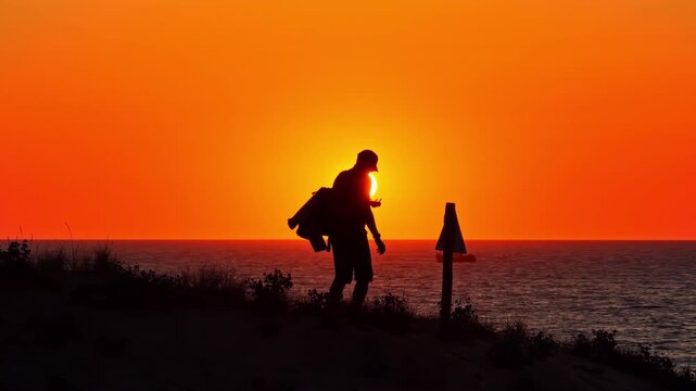 Backlit hiker stands on a dune at Plage des Oc&eacute;anides, silhouetted against a vivid orange sea sunset with a distant boat.
