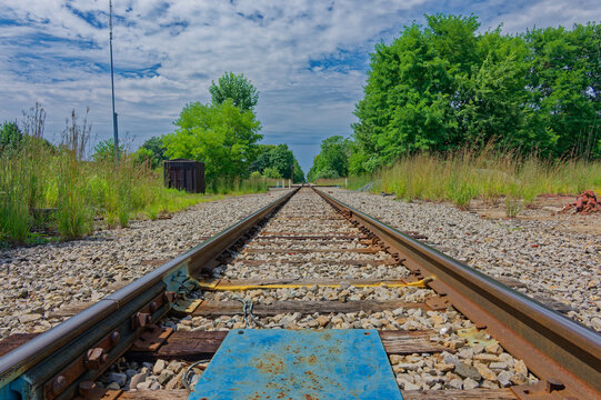 Low-angle view of railroad tracks stretching into a lush green countryside, emphasizing perspective, vanishing point and textured wooden ties with gravel; calm, rural atmosphere and open sky. in Plymo