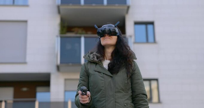 Young woman in a parka flies an FPV drone using goggles and a hand controller in front of modern apartment buildings.