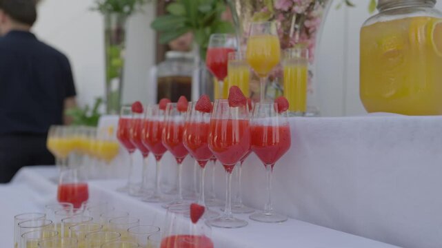 Rows of strawberry cocktails garnished with fruit beside orange juice glasses on a white table
