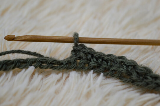 Wooden crochet hook inserted into a row of green wool yarn stitches resting on a white fluffy fur background close-up. Crocheting project, handmade yarn craft, needlework process, and cozy hobby conce