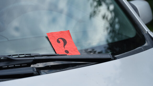 A crumpled red paper note with a black question mark is placed under a windshield wiper of a car, representing a mystery message or an unexpected notification.
