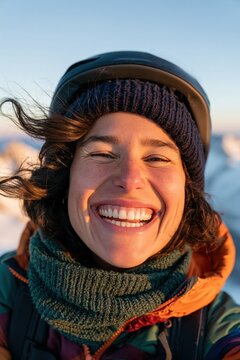 Smiling woman in winter hiking gear wearing a helmet and knit beanie with windblown hair and snowy mountains blurred in the background.