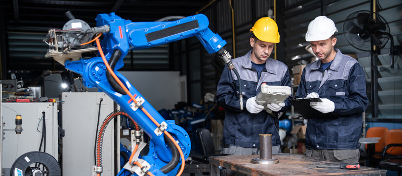 Factory technicians verify automated welding robot program code using hand controller and paper report clipboard