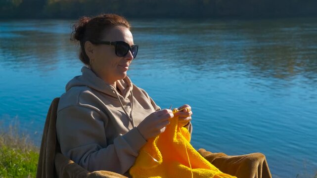 Woman knitting outdoors relaxing by a river. Peaceful middle-aged woman wearing sunglasses and a hoodie enjoying her knitting hobby while sitting in a chair by the river on a sunny day