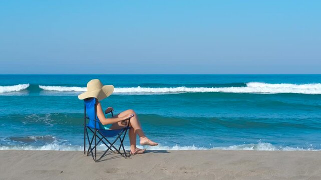 Woman relaxing on beach chair watching ocean waves. Sunlit tourist relaxing on beach chair, wearing straw hat and swimsuit, gazing at ocean waves washing over sandy shoreline during tropical vacation