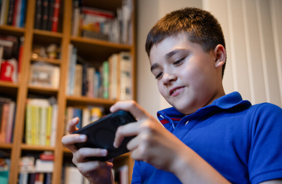 Happy Young Boy Using Mobile Phone While Sitting In Study Room, Child Holding Smartphone In Landscape Mode For Gaming, Online Entertainment, Digital Technology, Internet And Screen Time At Home.