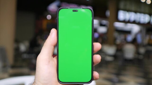 Hand holds a smartphone with a green screen. Blurred background shows a busy shopping mall. People move casually with neutral expressions. The green screen suits app demonstrations