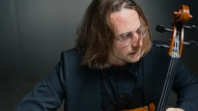 Young adult Caucasian man with glasses playing cello, camera tilting down from shaking hair to hands bowing strings. Footage is suitable for music school promos or classical concert ads