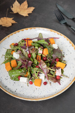 Plate with salad with greens, pumpkin and soft cheese and cutlery on dark background.