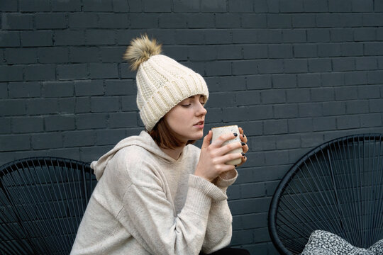 A young teenage woman with a mug of warm drink in her hands in a sweater and a beanie hat