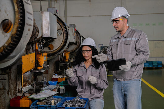 Experienced male industrial engineer wearing a hard hat and safety glasses carefully instructing a female worker on how to safely operate a large heavy machinery press inside manufacturing factory.