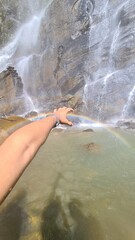 Obraz premium A person's hand reaches out to touch a beautiful rainbow forming in the mist of a powerful waterfall cascading down rocky cliffs into a clear pool of water