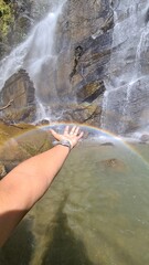 Obraz premium A hand reaching towards the vibrant spectrum of a rainbow, gracefully arcing over the tranquil waters at the base of a powerful waterfall, a serene scene of natural beauty and wonder