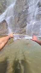Obraz premium Person holding a vibrant rainbow between their hands in front of a majestic mountain waterfall cascading into a clear pool