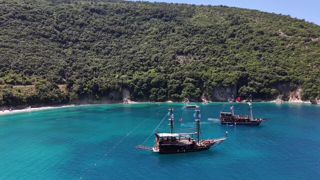 Several excursion boats with masts sit in turquoise water at Kakome bay, Albania, beneath a densely vegetated hillside. A small dinghy is visible in the foreground.
