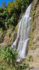 Scenic mountain waterfall cascading down rocky cliff with lush greenery and forest foliage © MdAbu