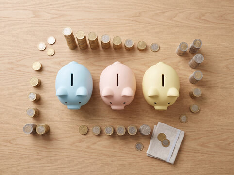 Three Pastel Piggy Banks with Coin Stacks on Wooden Desk, Flat Lay
